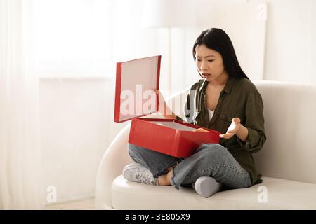 Enttäuschte asiatische Frau, die den roten Karton auspackte und zu Hause mit überkreuzten Beinen auf dem Sofa saß Stockfoto