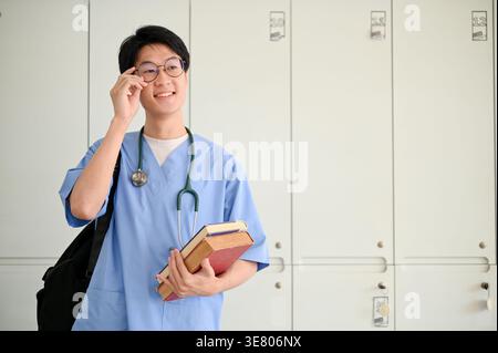 Lächelnder asiatischer männlicher Medizinstudent, der eine Brille mit Büchern in studentâ Umkleideraum justiert Stockfoto