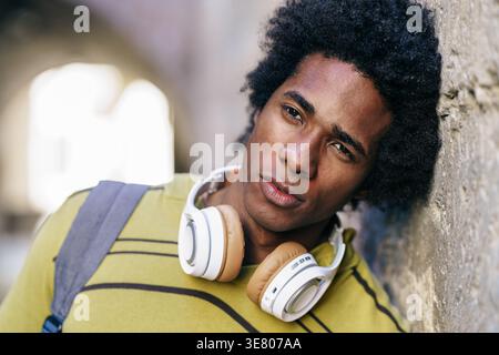 Seriöser Schwarzer mit Afro-Kopfhörern um den Hals, der draußen an die Betonwand gelehnt ist Stockfoto