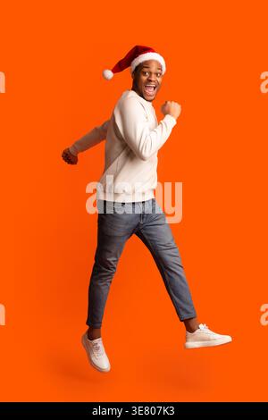 Schwarzer Mann in weißem Pullover und Weihnachtsmannmütze, der Spaß hat, im Studio gegen orangen Hintergrund zu springen Stockfoto