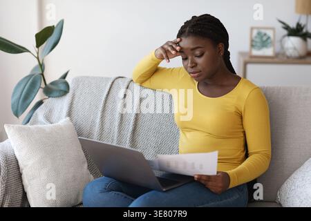 Verärgerte Schwarze Frau liest Dokumente, die den Kopf berühren, während sie auf dem Sofa sitzt und im Wohnzimmer zu Hause mit Laptop auf dem Schoß sitzt Stockfoto