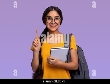 Porträt einer indischen Schülerin mit Brille und Rucksack, die Ordner und Bleistift hält, während sie vor violettem Hintergrund steht Stockfoto