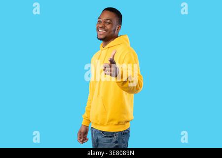 Lächelnder bärtiger Schwarzer Mann mit gelbem Hoodie, der den Finger nach vorne zeigt, während er seitlich vor blauem Hintergrund im Studio steht Stockfoto