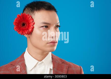 Seriöser asiatischer Mann, der Augen-Make-up in einem karierten roten Anzug mit roter Blume hinter dem Ohr trägt und vor blauem Hintergrund im Studio steht Stockfoto