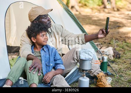 Ein schwarzer Mann mittleren Alters, der ein Lifestylephone hält und Selfie mit Enkel macht, während er während des Campingens im Wald auf dem Zelt sitzt Stockfoto