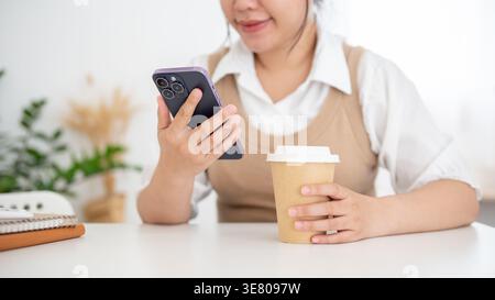Zugeschnittenes Bild einer asiatischen Frau, die eine Kaffeetasse hält, mit einem Smartphone, das zu Hause am Tisch sitzt Stockfoto