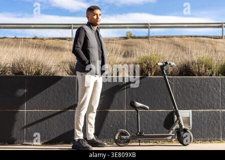 Hispanischer Mann mit Sweatshirt und ärmelloser Pufferjacke, der an sonnigem Tag seitlich in der Nähe eines elektrischen Rollers steht Stockfoto