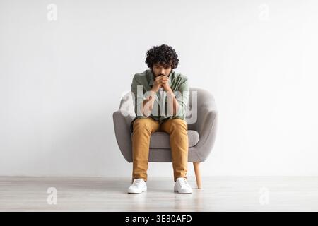 Asiatischer Mann mit lockigen Haaren, das Kinn auf gefalteten Händen stützt und wegschaut, während er auf dem Stuhl vor der weißen Wand zu Hause sitzt Stockfoto