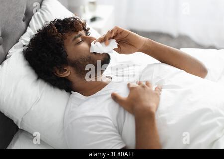 Asiatischer Mann mit lockigen Haaren, der die Nase mit einem Taschentuch abwischt, während er mit geschlossenen Augen auf dem Bett zu Hause im Schlafzimmer liegt Stockfoto