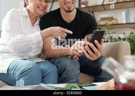 Lächelnder gemischter Sohn und ältere Mutter beobachten auf dem Smartphone, während sie zusammen auf dem Sofa im Wohnzimmer sitzen Stockfoto