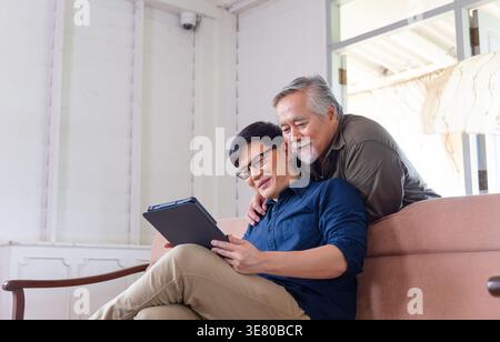 Asiatischer älterer Vater, der sich hinter seinem Sohn mittleren Alters mit einer Brille in der Hand eines Tablets auf der Couch zu Hause liegend Stockfoto