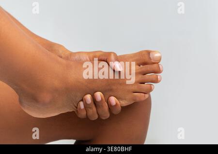 Schwarze Frau, die unter Fußschmerzen leidet, massiert ihren Fuß vor grauem Hintergrund im Studio Stockfoto