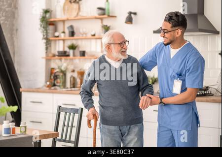 Lächelnder arabischer männlicher Betreuer in Brille, der einen älteren Mann im Stehen zu Hause ansieht Stockfoto
