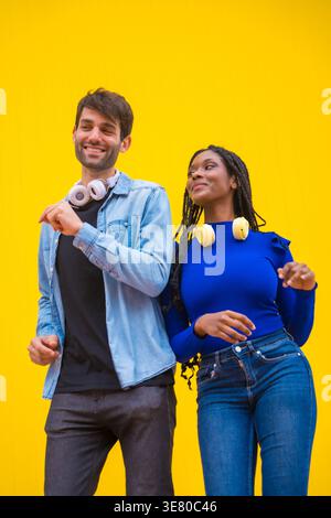 Lächelnde Schwarze und kaukasische Paare mit Kopfhörern um den Hals tanzen zusammen vor gelbem Hintergrund im Studio Stockfoto