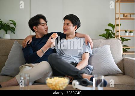 Lächelnder asiatischer schwuler Mann, der seinen Freund umarmt und ihm einen Donut füttert, während er im Wohnzimmer zu Hause dicht beieinander sitzt Stockfoto
