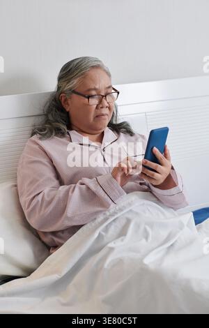 Asiatische ältere Frau mit Brille mit Smartphone, während sie im Bett im Krankenhauszimmer sitzt Stockfoto