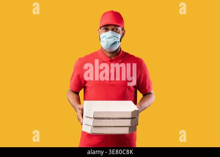 Schwarzer Mann mit Hut und Gesichtsmaske, der Pizzakästen hält, während er im Studio vor gelbem Hintergrund steht Stockfoto