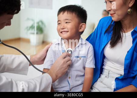 Gemischter Junge, der einen männlichen kaukasischen Arzt ansieht, wie er seinen Herzschlag hört und mit Mutter in der Klinik sitzt Stockfoto