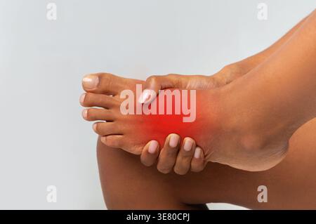 Schwarze Frau, die unter Fußschmerzen leidet, massiert ihren Fuß vor grauem Hintergrund im Studio Stockfoto
