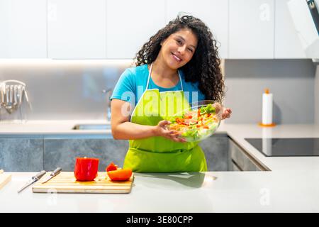 Hispanische Frau mit lockigem Haar, die grüne Schürze trägt und eine Schüssel mit Gemüsesalat hält, während sie zu Hause an der Küchenzeile steht Stockfoto