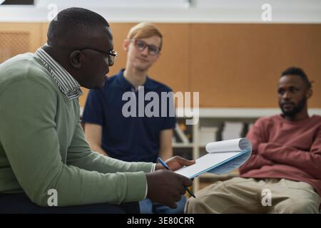 Seitenansicht eines schwarzen männlichen Psychologen, der eine Brille trägt und das Klemmbrett hält und mit schwarzen und kaukasischen männlichen Patienten spricht, während er auf dem Stuhl sitzt... Stockfoto