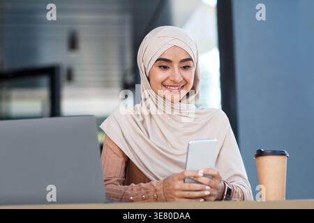 Lächelnde indische muslimische Frau im Hijab mit Smartphone, die tagsüber am Tisch mit Laptop im Café im Freien sitzt Stockfoto