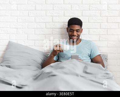 Lächelnder Schwarzer Mann, der sein Smartphone benutzt und Kaffee trinkt, während er zu Hause auf dem Bett sitzt Stockfoto