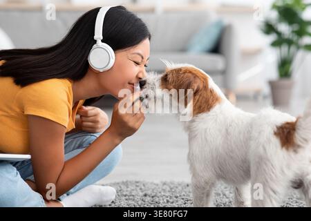Asiatische Frau mit Kopfhörern kuschelt Hund auf dem Boden mit Teppich im Wohnzimmer zu Hause Stockfoto