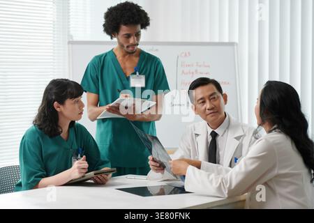 Schwarze männliche und kaukasische Assistenzärzte sitzen am Tisch und hören asiatischen Ärzten zu, wie sie das Röntgenergebnis im Konferenzraum in der Klinik besprechen Stockfoto