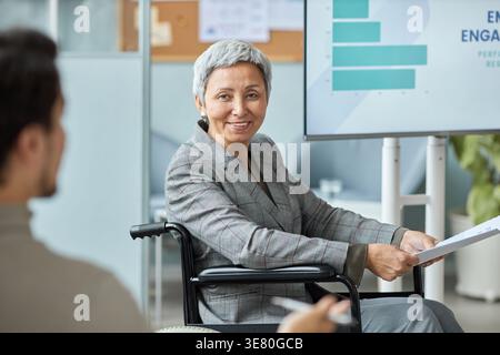 Leitende asiatische Geschäftsfrau, die im Rollstuhl sitzt und während der Besprechung im Büro eine Präsentation hält Stockfoto