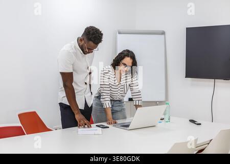 Schwarzer Mann und weiße Frau mit Brille, die am Tisch stehen und sich über einen offenen Laptop beugen, um im Konferenzraum über ein Projekt zu diskutieren Stockfoto