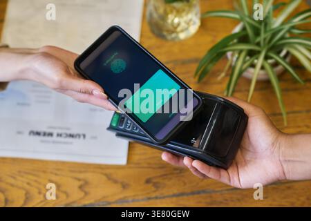 Schwarze weibliche Mitarbeiterin mit POS-Terminal, die Zahlung von einer kaukasischen Kundin per Smartphone über dem Tisch im Café erhält Stockfoto