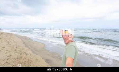 Hispanischer Mann mit Mütze und Sonnenbrille, der Selfie macht, während er an sonnigen Tagen am Strand steht Stockfoto