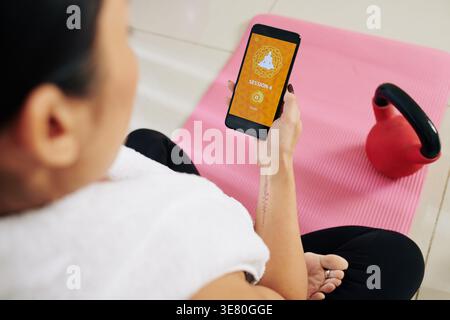 Asiatische Frau, die fünf Minuten Yoga-Sitzung auf dem Smartphone beginnt, während sie auf der Yoga-Matte zu Hause sitzt Stockfoto