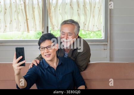 Asiatischer älterer Vater, der hinter einem Mann mittleren Alters steht und ein Smartphone benutzt, das auf der Couch sitzt, während beide im Wohnzimmer zu Hause Videoanrufe führen Stockfoto