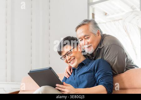 Asiatischer älterer Vater, der sich hinter seinem Sohn mittleren Alters mit einer Brille in der Hand eines Tablets auf der Couch zu Hause liegend Stockfoto
