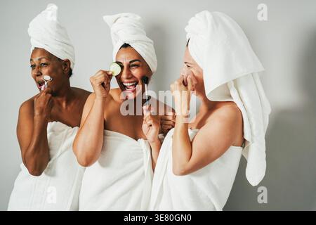 Verschiedene Frauen, die Badetücher tragen, haben Spaß mit Hautpflegeprodukten, während sie im Studio vor grauem Hintergrund stehen Stockfoto