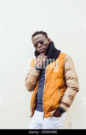 Schwarzer Mann, der Jacke mit Schal und Sonnenbrille trägt und mit Hand in der Tasche vor der weißen Wand steht Stockfoto