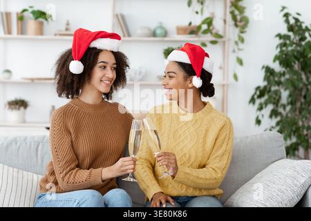 Lächeln verschiedene weibliche Freunde mit Weihnachtsmützen und Pullover, die Gläser mit Champagner auf dem Sofa zu Hause im Wohnzimmer tosten Stockfoto