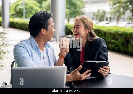 Fröhliche thailändische Geschäftskollegen feiern gute Nachrichten, indem sie Siegesfäuste zeigen, die mit Laptop und Tablet im Café am Tisch sitzen Stockfoto