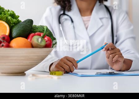 Zugeschnittenes Bild einer schwarzen Ernährungsberaterin, die einen Stift hält, der mit einer Schale Obst und Gemüse in der Klinik am Tisch sitzt Stockfoto