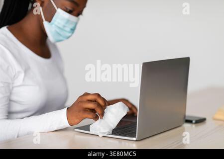 Schwarze Frau mit Zöpfen, die eine medizinische Maske trägt, die ihren Laptop reinigt, während sie am Tisch in der Küche zu Hause sitzt Stockfoto