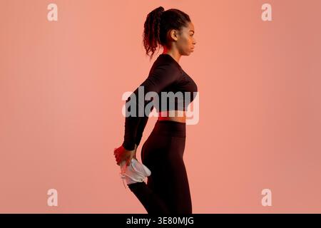 Schwarze Frau mit Sportbekleidung, das Bein vor rosa Hintergrund im Studio dehnt Stockfoto