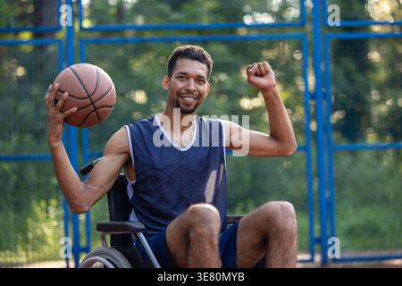 Schwarzer Mann mit Behinderung, der tagsüber im Rollstuhl auf einem Basketballfeld sitzt Stockfoto