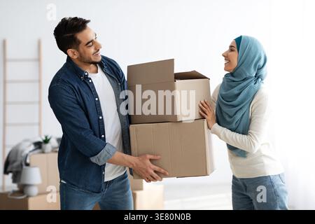 Mann aus dem Nahen Osten und muslimische Frau im Hijab stehen und tragen Papierkästen zusammen, während sie in ihr neues Haus ziehen Stockfoto
