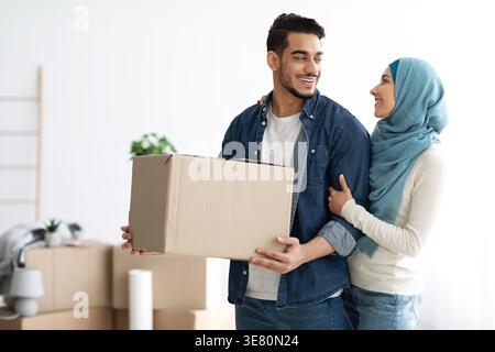 Ein Mann aus dem Nahen Osten, der eine Kiste trägt und seine muslimische Frau im Hijab ansieht, der seinen Arm umschließt, während er in ihr neues Zuhause zieht Stockfoto