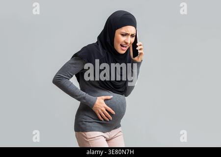 Ängstliche arabische schwangere Frau, die Hijab trägt, ihren Bauch berührt und einen Anruf hat, während sie vor grauem Hintergrund steht Stockfoto