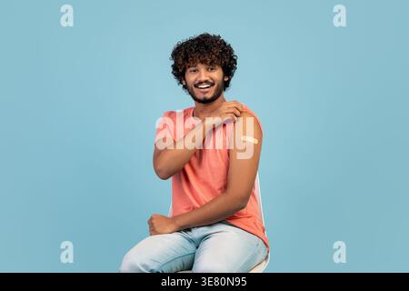 Bärtiger indischer Mann mit lockigen Haaren, der einen geimpften Arm zeigt, während er auf Stuhl vor blauem Hintergrund im Studio sitzt Stockfoto