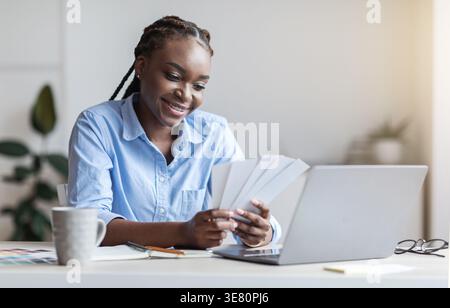 Schwarze Geschäftsfrau mit Zöpfen, die Farbpalette halten, während sie im Heimbüro mit Laptop am Tisch sitzt Stockfoto