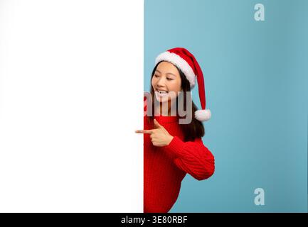 Asiatische Frau mit Weihnachtsmann-Hut, der mit dem Finger zeigt und hinter weißem Banner vor blauem Hintergrund im Studio steht Stockfoto